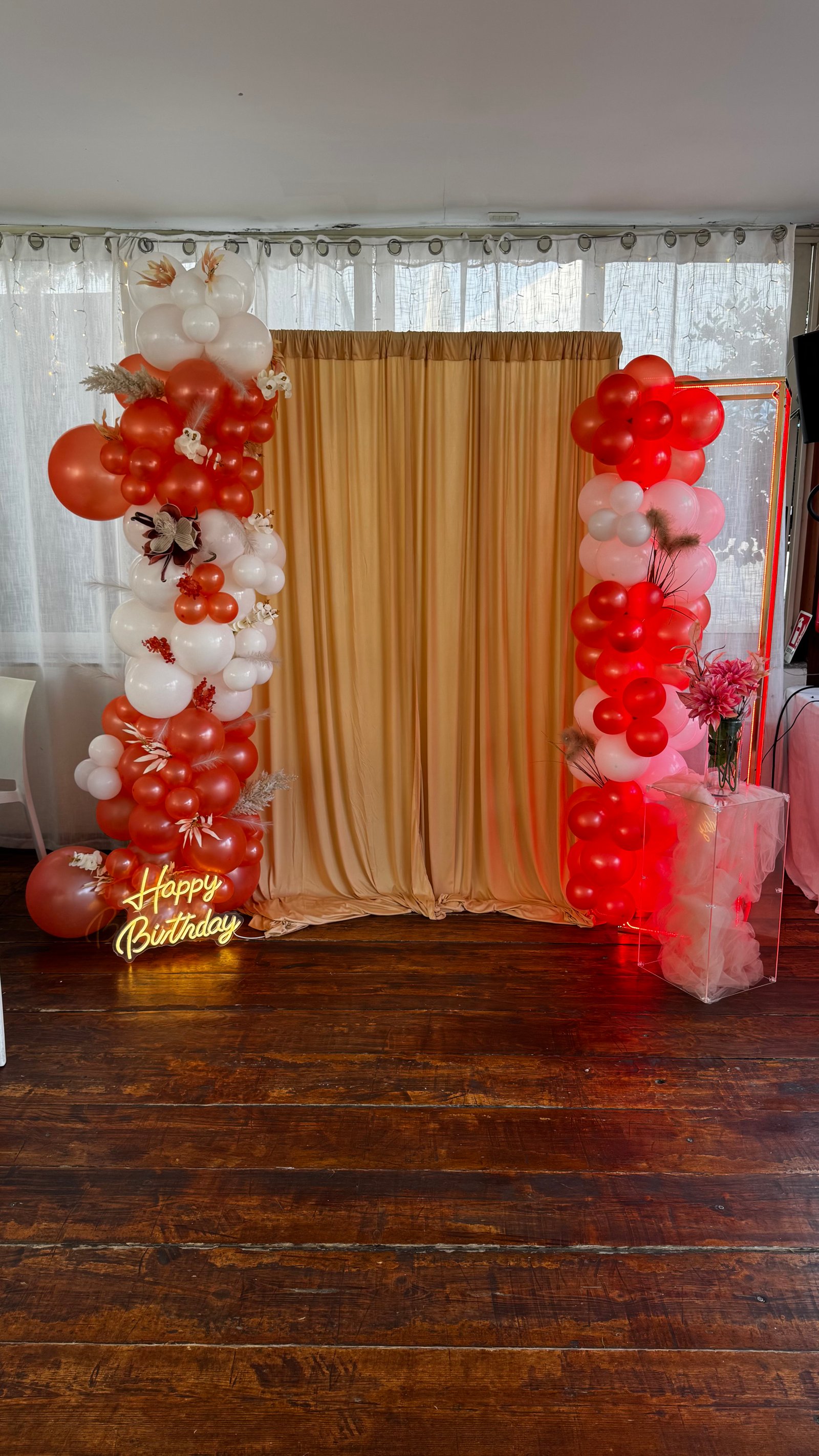 Allestimento compleanno con palloncini arancioni, bianchi e rossi, tenda beige e neon Happy Birthday